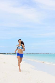 Beach running Asian woman doing morning cardio workout on white sand and turquoise ocean water training cardio for weight loss. Copyspace on blue sky.