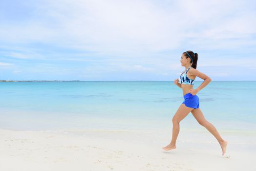 Healthy and active lifestyle running woman jogging on beach. Full length young female adult doing morning cardio workout barefoot in white sand and turquoise ocean background.