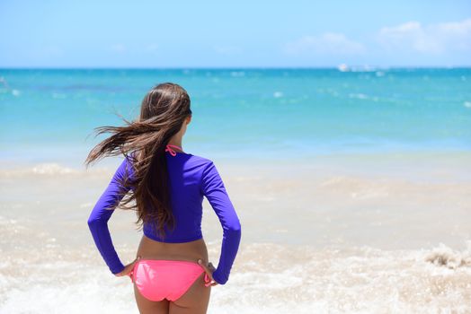 Beach girl going swimming in rashguard swimwear protective clothing. Woman standing in bikini and uv sun protection surf shirt looking at ocean waves. Healthy active lifestyle.