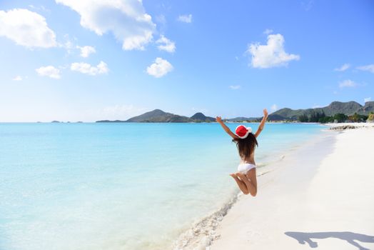 Christmas beach travel vacation woman in Santa hat jumping of joy on tropical Caribbean winter holidays getaway. Beautiful girl in bikini having fun under the sun traveling.