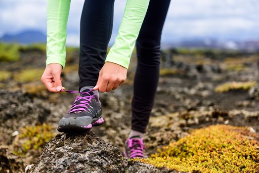 Trail runner woman getting ready to run on rocky mountain mud race in autumn. Athlete tying up laces of running shoes. Closeup of hands.