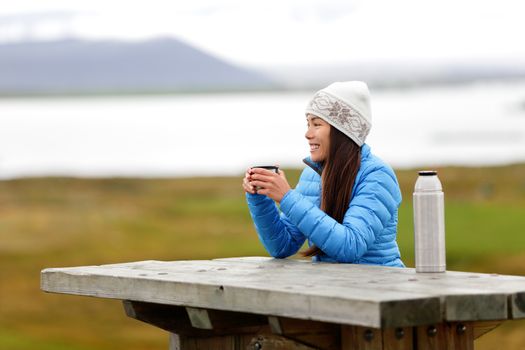 Woman in outdoors drinking coffee from thermos bottle flask sitting outside wearing warm down jacket and knit hat. Active young mixed race Asian Chinese Caucasian woman enjoying traveling on Iceland.