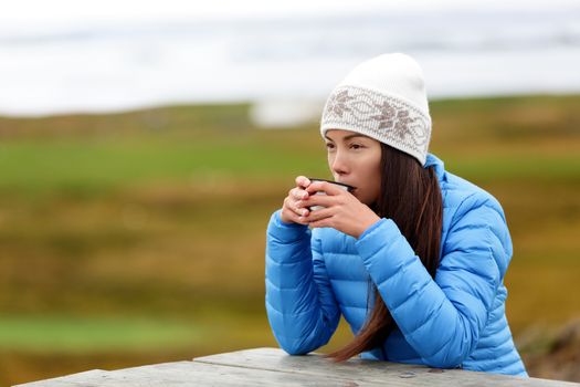 Woman in outdoors drinking coffee from thermos cup sitting outside wearing warm down jacket and knit hat. Pretty young mixed race Asian Chinese Caucasian woman living active lifestyle.