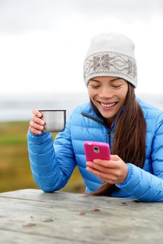 Woman outdoors using smart phone app drinking coffee from thermos cup sitting outside wearing warm down jacket and knit hat. Pretty young mixed race Asian Chinese Caucasian woman in active lifestyle.