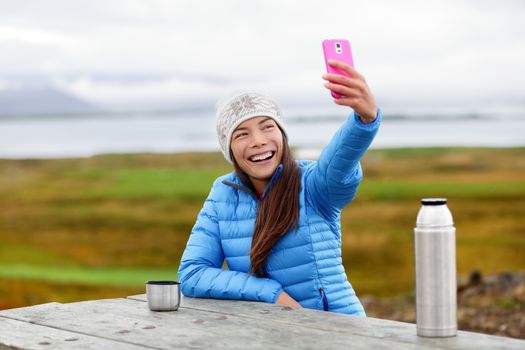 Woman outdoors using smart phone taking selfie photo on app on smartphone while sitting outside wearing warm down jacket. Pretty young mixed race Asian Chinese Caucasian woman in active lifestyle.