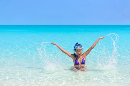 Beach holiday woman playing in the ocean. Asian young adult wearing a snorkel scuba mask having fun splashing water with arms up and swimming in vacation resort travel destination.