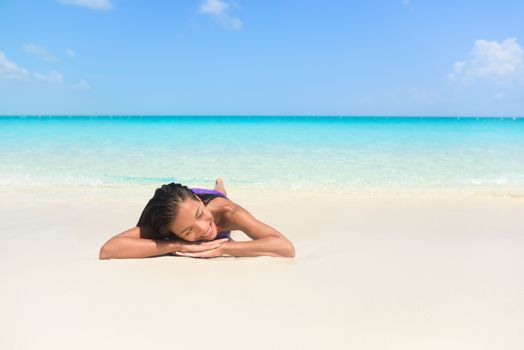 Relaxing woman on beach vacation sleeping on sand. Beautiful girl lying down under the sun tanning in perfect paradise white sand beach and pristine blue ocean background.