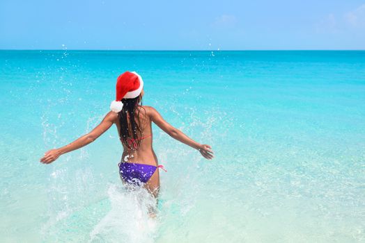 Christmas beach bikini woman swimming in ocean. Beautiful adult entering perfect turquoise water with arms back in freedom feeling free enjoying winter vacations wearing a santa hat for the holidays.