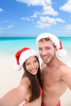 Happy couple on Christmas travel holidays taking selfie picture with smartphone wearing santa hat during their winter vacation. Young adult friends in swimsuit and bikini in front of the ocean.