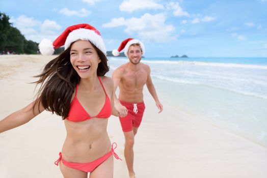 Happy Christmas couple having fun running on beach. Beautiful young Asian woman and Caucasian boyfriend holding hands playing on sand during winter travel holidays wearing santa hats.