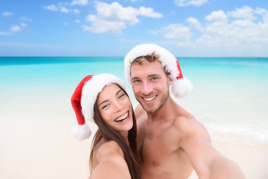 Christmas couple selfie picture on beach vacation. Happy young adults smiling at camera taking self-portrait wearing santa hats. Multiracial Caucasian and Asian people.