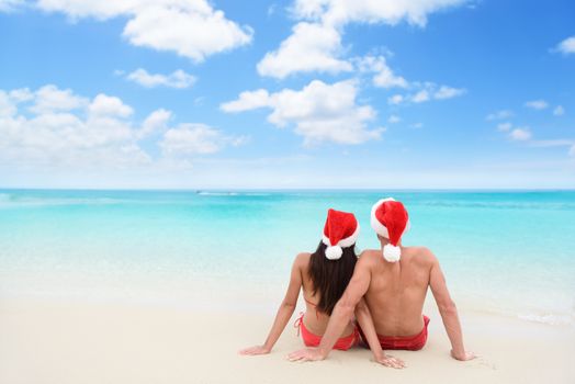 Christmas couple in love lying down relaxing on white sand beach sun tanning in tropical travel destination during winter holidays. Back view of young adults wearing santa hat.
