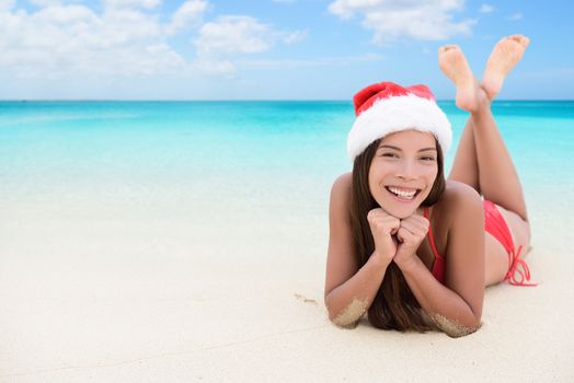 Christmas woman relaxing during winter beach holidays. Girl smiling at camera during travel vacation wearing a santa hat. Young mixed race adult in her 20s.