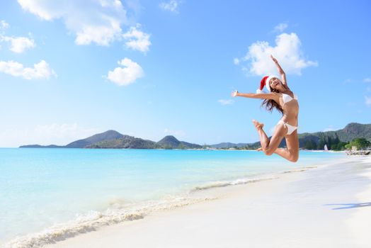 Happy Christmas vacation - girl jumping of joy and surprise on perfect white sand beach for winter holidays. Young woman wearing santa hat arms raised of happiness during vacations.