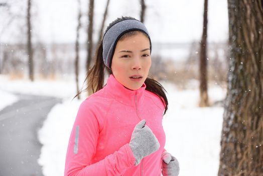Woman running in cold weather wearing winter accessories, pink windbreaker, gloves and headband. Asian Chinese young adult doing her cardio exercises outside in a city park.