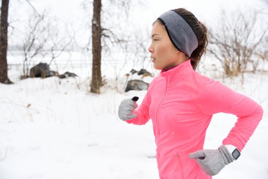 Female runner trail running in cold snowing weather. Asian Chinese athlete woman training for marathon jogging outside in snow wearing activewear jacket, headband, and winter gloves.