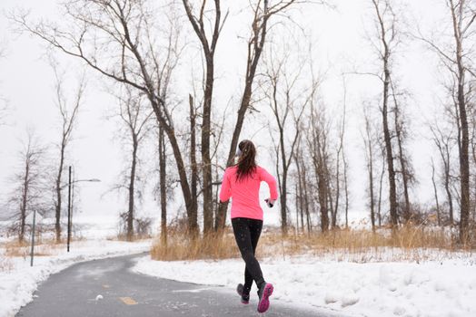 Woman runner running in winter snow - active lifestyle. Female athlete from the back jogging training her cardio on city park path outside in cold weather wearing leggings and coat.