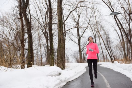 Woman jogging in snowy city park - winter fitness. Female athlete exercising outside in cold weather on forest path wearing activewear. Windbreaker pink jacket, warm tights, running shoes.