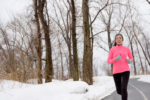 Winter cardio exercise - woman jogging doing her workout outside. Young adult running in outdoor park with snowy forest background wearing cold weather gear.