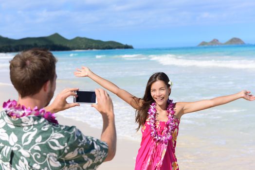 Man taking pictures with smart phone of woman on beach on Hawaii. Young couple having fun living happy lifestyle on Hawaiian beach holiday vacation travel. Man using smartphone for photo.