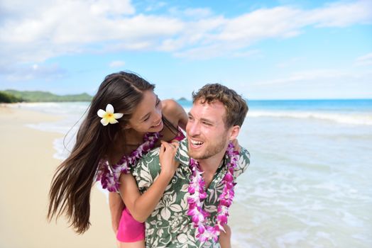 Beach couple having fun laughing on Hawaii holiday. Beautiful Asian mixed race woman piggybacking on Caucasian boyfriend wearing traditional Hawaiian lei on Oahu, Hawaii, USA.