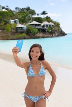 Beach vacation travel woman making smart phone selfie in bikini having fun sharing on social media. Self portrait photo with smartphone on beach. Happy mixed race Caucasian / Asian Chinese woman.
