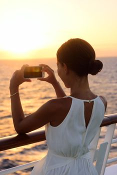 Cruise ship vacation woman taking photo with smart phone camera enjoying sunset on travel at sea. Girl using smartphone to take picture of ocean sunset. Woman in dress on luxury cruise liner boat.