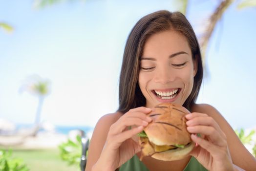 Happy woman holding eating fresh burger sandwich at beach. Young female is sitting at outdoor restaurant. Beautiful tourist is having fresh burger during summer vacation holidays.