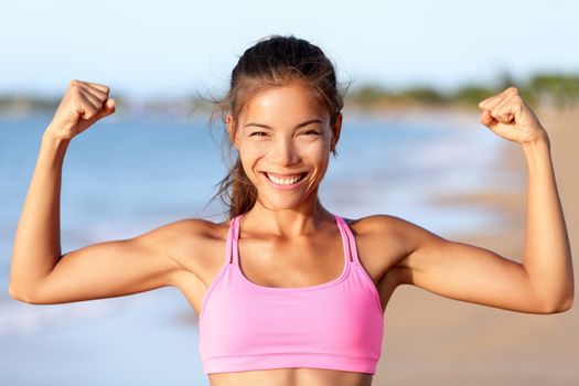 Happy sporty fitness woman flexing muscles on beach. Smiling young is wearing pink sports bra. Female is showing her strength and healthy lifestyle on sunny day.