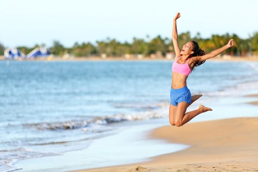 Excited woman jumping at beach. Cheerful female in sportswear is enjoying on shore. Runner with arms raised screaming in midair on sunny day.