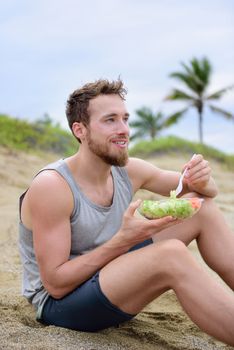 Fitness man eating healthy salad meal at workout. Handsome young muscular male adult sitting on the beach after running workout for lunch break with fresh prepared to go vegan raw vegetables food.