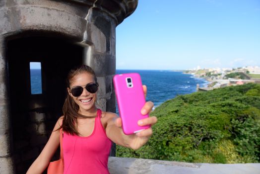 Tourist taking fun selfie at famous landmark. Travel woman holding a pink smartphone visiting Old San Juan's Castillo San Felipe Del Morro, the main attraction of the city of San Juan, Puerto Rico.