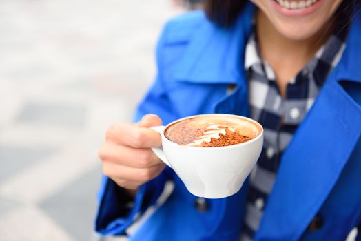 Hands closeup of woman drinking at cafe holding a coffee cup with a milk rosetta foam shape on top. Outdoor terrace at coffee shop or restaurant.