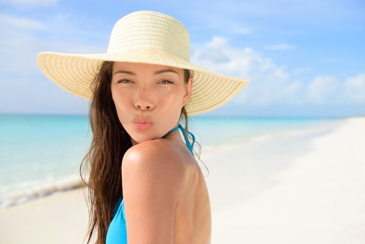 Beach sun hat woman blowing cute kiss on vacation. Asian female young adult model striking a kissing pose to the camera for summer holidays wearing straw sunhat and blue bikini on perfect white sand.