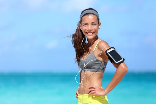 Happy fitness woman living a fit healthy lifestyle. Young Asian Caucasian girl wearing activewear and sports armband for phone and earphones, tech gear for running or cardio workout on beach.