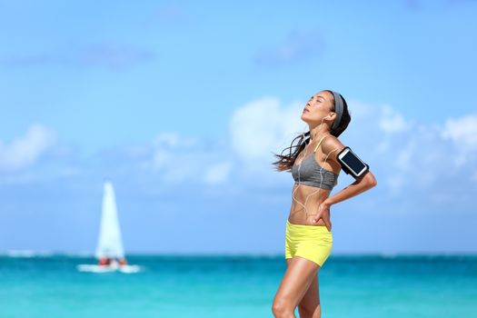 Tired jogging woman catching her breath taking a break of hard workout. Young Asian female athlete runner holding her back in pain after intense cardio running exercise on long beach run.