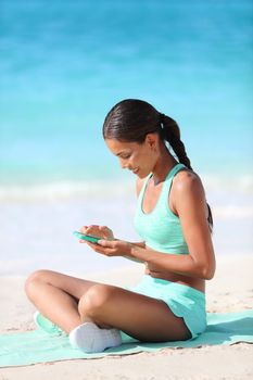 Fit girl using fitness app on phone during travel holidays on beach texting or posting on social media online. Healthy sporty Asian woman living a happy active life exercising on towel.