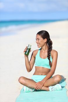 Fitness woman drinking a healthy green smoothie juice for a detox weight loss cleanse. Happy sporty asian girl on beach taking a snack break with a vegan beverage for a vegetarian diet.