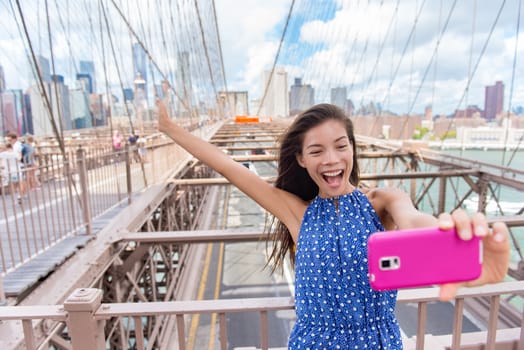 Happy selfie tourist woman taking fun self-portrait picture with smart phone app on Brooklyn Brige, New York City, Manhattan, USA. Asian woman posing with smartphone for social media.