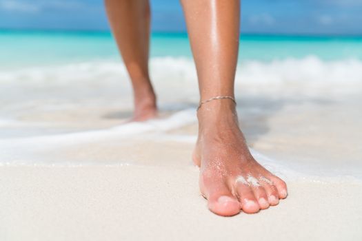 Beach feet closeup - barefoot woman walking in ocean water waves. Female young adult legs and toes wearing an ankle bracelet anklet relaxing in summer vacation travel.