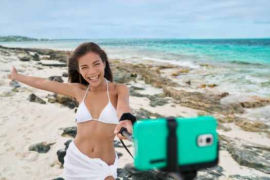 Holiday vacation girl taking smart phone self portrait pictures with selfie stick. Asian woman having fun doing mobile photography with smartphone for social media on caribbean beach summer travel.