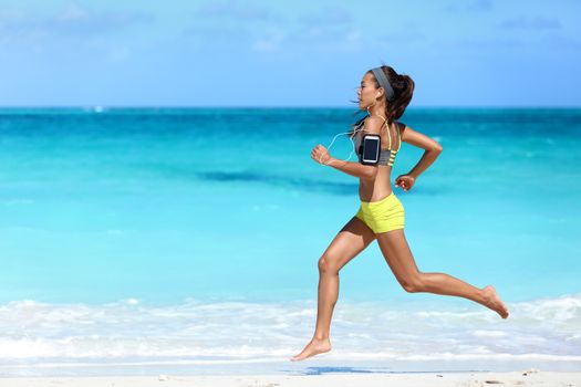 Fitness runner woman running on beach listening to music motivation with phone case sport armband strap. Sporty athlete training cardio barefoot with determination under summer sun.