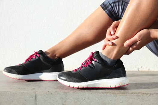 Ankle jogging injury female runner holding painful sprained ankle. Legs and running shoes closeup of woman hands gripping on leg in pain because of sore and injured joint and muscles in ankles.