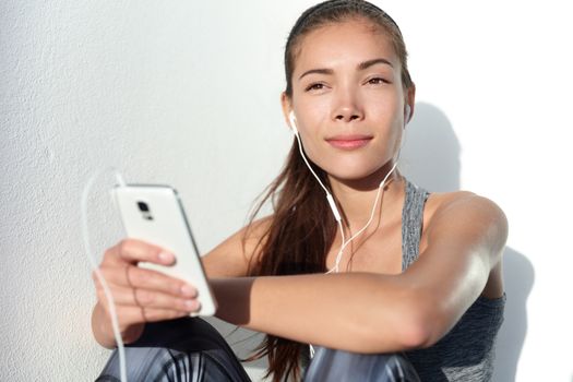 Young woman listening to music with earphones on smart phone app for fitness motivation. Athlete runner in sportswear relaxing sitting getting inspired. Asian mixed race model.