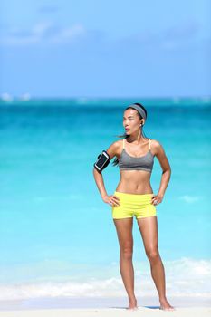 Fit slim body fitness woman runner getting ready for running listening to motivation music on phone with smartphone holder armband strap and earphones. Sexy athlete standing on beach in sports bra.