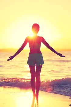 Mindfulness woman practicing yoga sun salutation at beach morning sunrise. Silhouette of fit person standing in sun flare raising arms to the sky with an open heart to do a meditation routine.