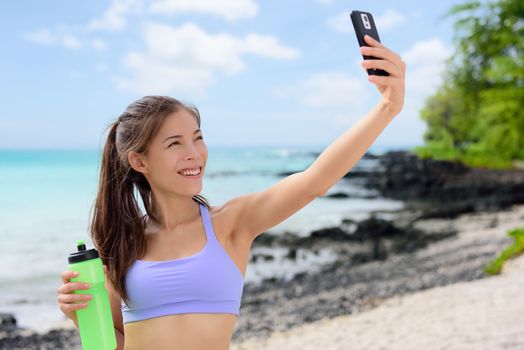 Smiling sporty woman taking selfie using smartphone on beach. Beautiful fit fitness female in sports bra is holding water bottle. Attractive young woman is posing at sea shore.