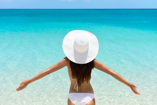 Woman on Beach standing with arms outstretched against turquoise sea. Rear view of female wearing white sunhat and bikini. Carefree tourist is enjoying vacation at beach.