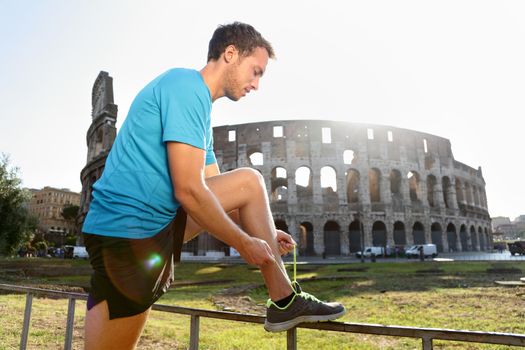 Young male jogger running tying shoelace against Colosseum. Side view of runner preparing for workout on sunny day. Athletic man is in sportswear.