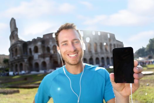 Happy young jogger showing smartphone with blank screen against Colosseum. Confident male runner is listening music on sunny day. The empty space on mobile phone can be used for advertisement.
